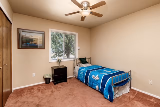 A small bedroom with beige walls and brown carpet featuring a single metal bed with blue bedding decorated with cartoon animals. Next to the bed is a black nightstand with a small vase of flowers and a trash can. A window with a view of trees is centered on the wall behind the bed, and a framed picture hangs on the adjacent wall. A ceiling fan with a light fixture is mounted on the ceiling.