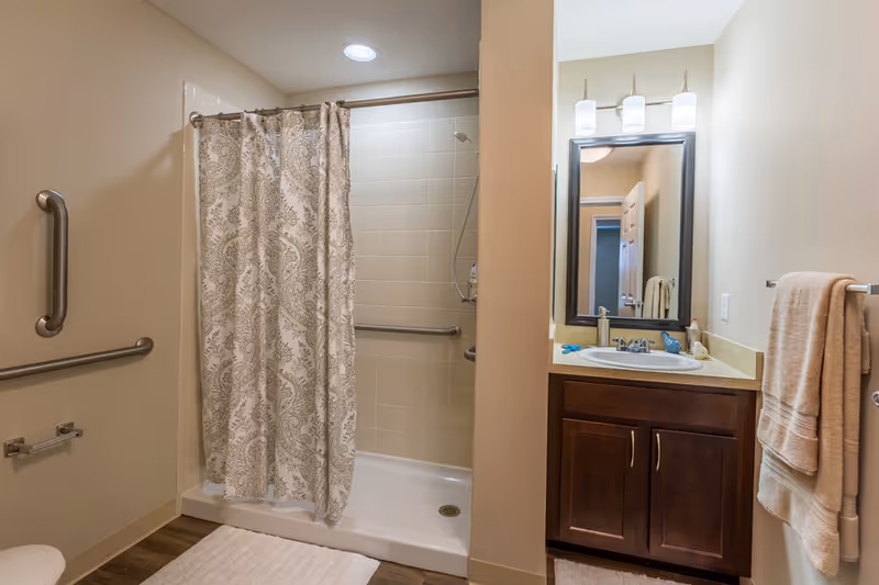 A bathroom with a walk-in shower featuring a patterned shower curtain and grab bars. There is a vanity with a sink, a mirror above it with three lights, and towels hanging on a rack on the wall. The floor has a white bath mat.