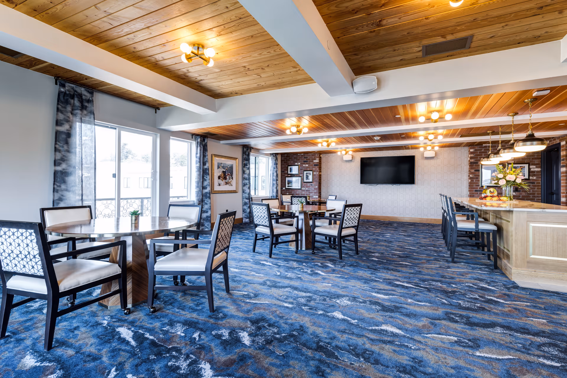 Spacious communal dining and lounge area with round tables and chairs, a bar counter, blue patterned carpet, and wood-paneled ceiling.