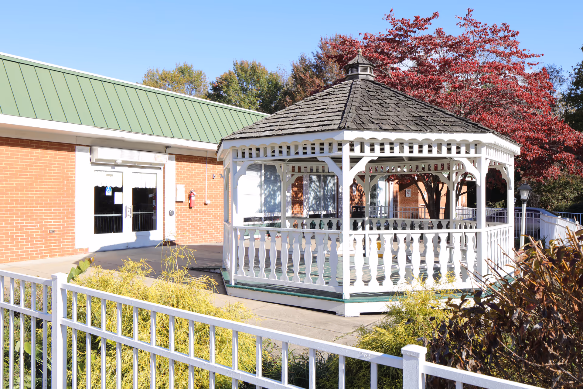 Outdoor view of a white wooden gazebo with a shingled roof situated on a concrete patio area next to a brick building with a green metal roof. There are trees with autumn foliage and some shrubs surrounding the area, along with a white metal fence in the foreground.