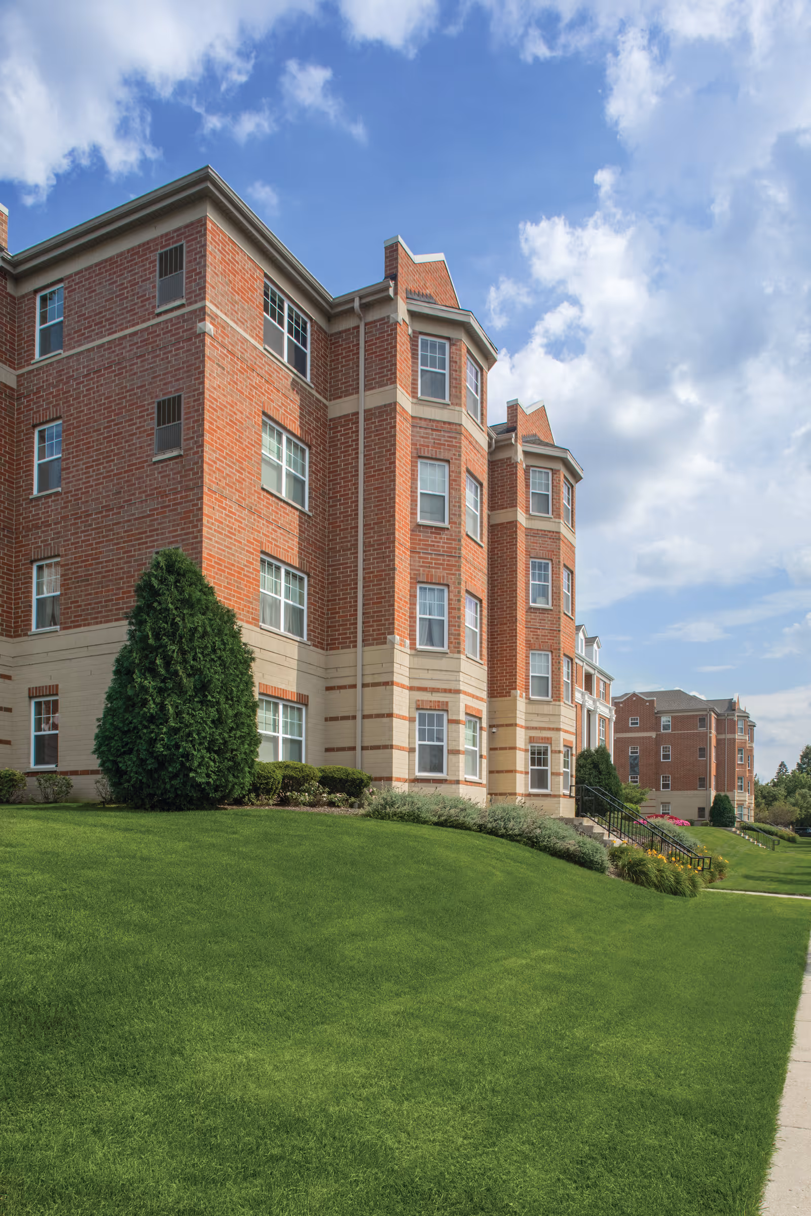 Exterior view of a multi-story brick building with beige accents, surrounded by well-maintained green grass and shrubs under a partly cloudy sky.