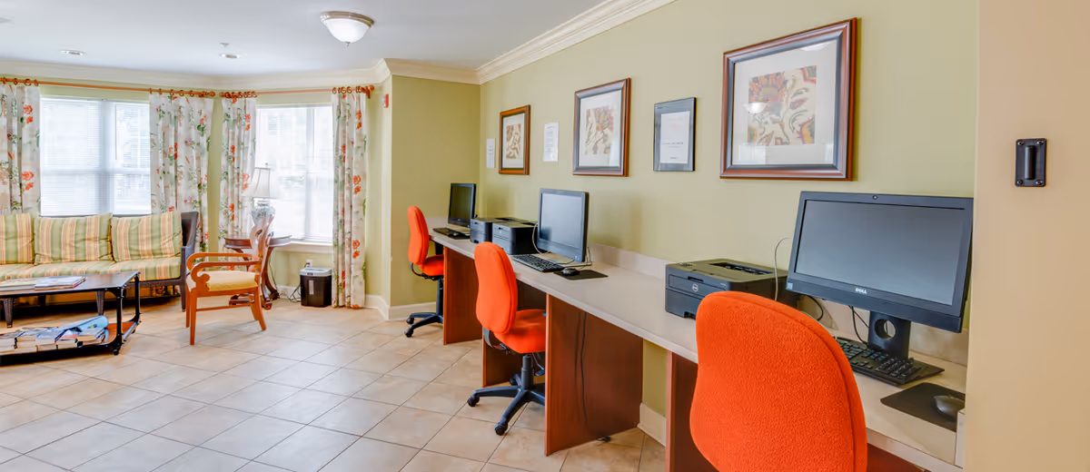 A senior living facility common area with a row of three computer workstations featuring desktop computers, keyboards, and printers. Each workstation has an orange swivel chair. To the left, there is a seating area with a striped sofa, wooden chairs, a coffee table, and floral curtains on large windows letting in natural light. The walls are painted light green and decorated with framed artwork.