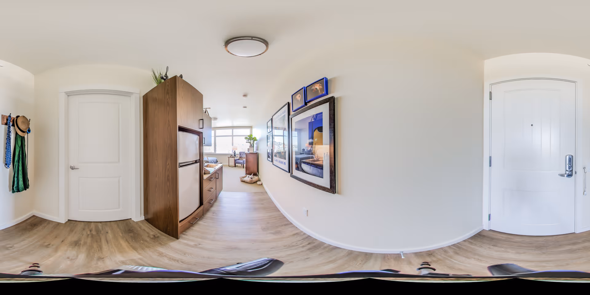 A bright and modern apartment interior with light wood flooring and white walls. On the left side, there is a white door and a wall-mounted coat rack with hats and scarves. In the center, a wooden cabinet with a small refrigerator is visible. The hallway leads to a living area with a large window letting in natural light, a sofa, a small table, and framed pictures on the right wall. The entrance door is on the right side of the image.