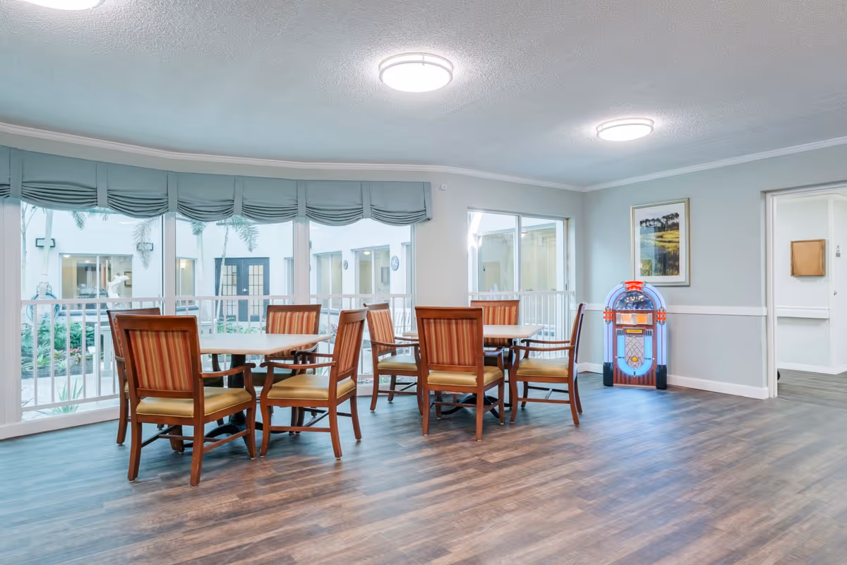 A bright and spacious common area with wooden tables and chairs arranged for seating. Large windows with blue valances allow natural light to fill the room, and a colorful jukebox stands against the wall. The flooring is wood-style, and the walls are painted light gray with white trim. A framed picture hangs on the wall.