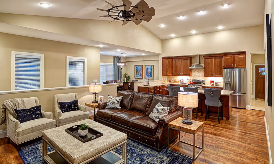 Open concept living room and kitchen area with a brown leather sofa, two beige armchairs, a large ottoman coffee table, and two side tables with lamps. The kitchen features wooden cabinets, stainless steel appliances, and a breakfast bar with two black bar stools. The room has wooden flooring, beige walls, and recessed lighting.