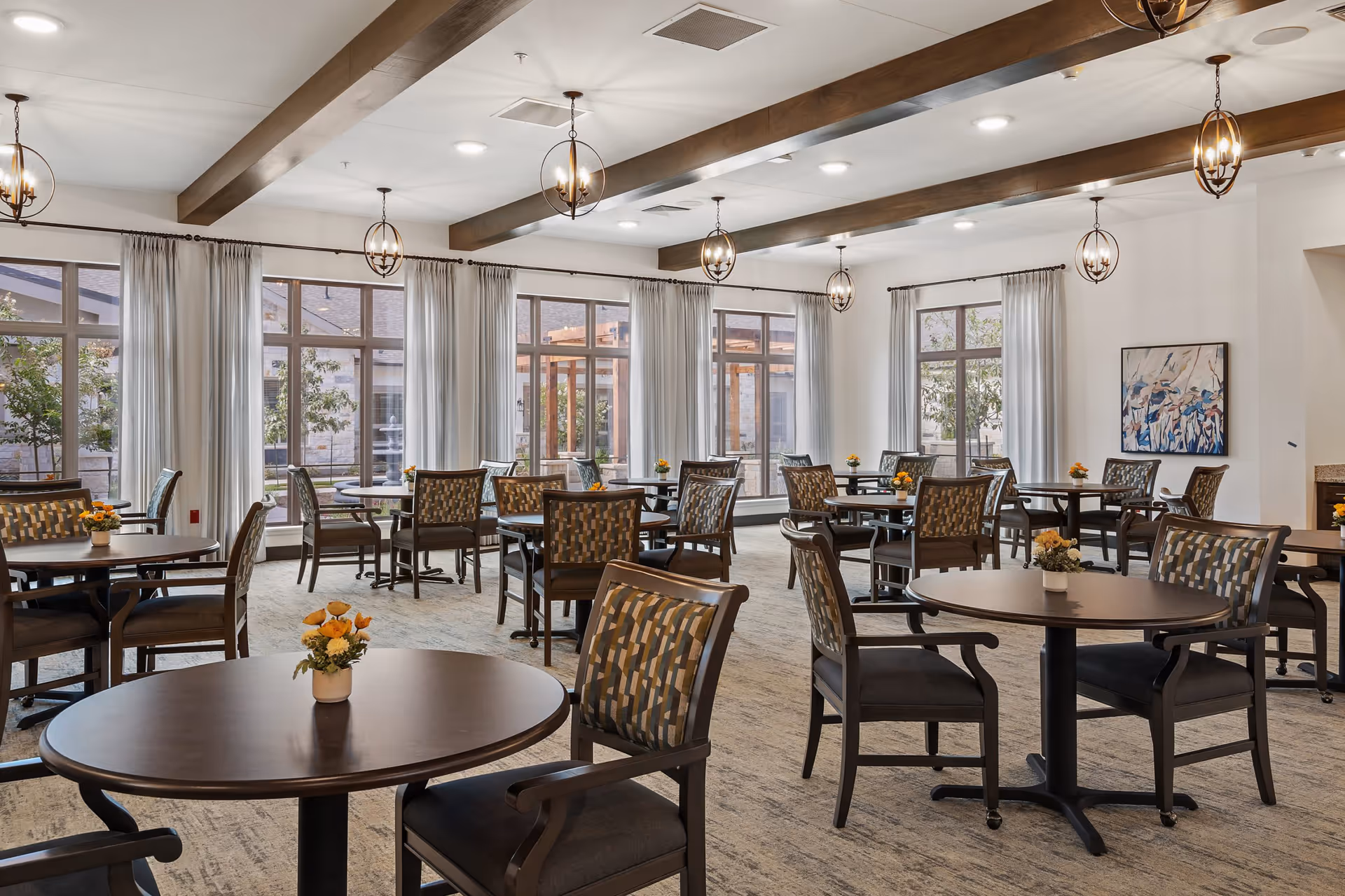 A spacious dining room with multiple round tables and cushioned chairs arranged neatly. Each table has a small floral centerpiece. Large windows with light curtains allow natural light to fill the room, and wooden ceiling beams with hanging light fixtures add warmth to the space. A painting is visible on one wall.