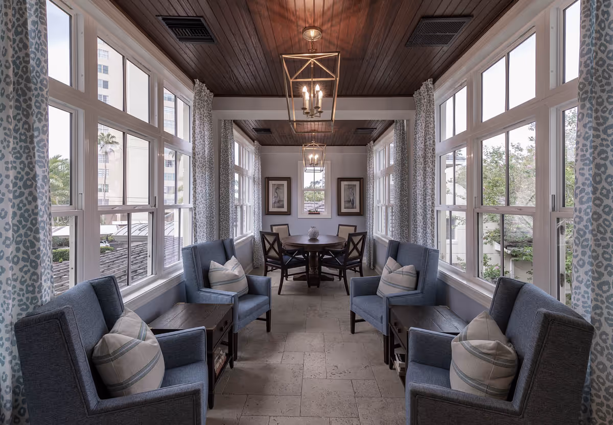 A bright and cozy sitting area with large windows on both sides, featuring four blue armchairs with striped pillows and two wooden side tables. At the end of the room, there is a round wooden table with four chairs. The ceiling is wooden with two modern pendant lights hanging down. Light blue patterned curtains frame the windows.