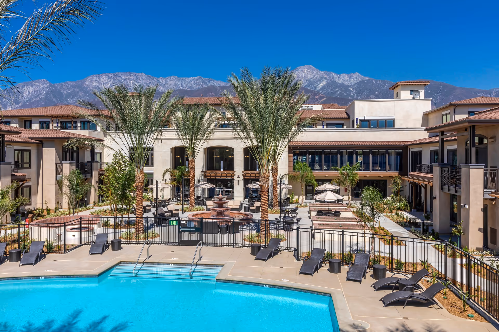 Outdoor view of Merrill Gardens at Rancho Cucamonga featuring a swimming pool with lounge chairs around it, palm trees, a central fountain, patio seating with umbrellas, and a multi-story building with balconies in the background under a clear blue sky and mountains.