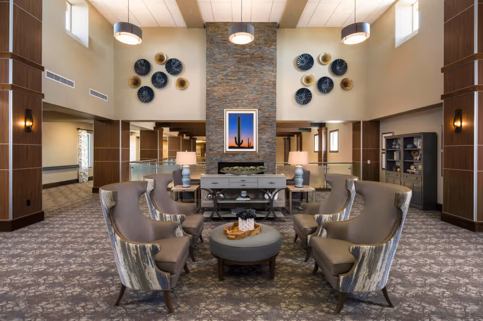 A spacious and elegant common area with six upholstered armchairs arranged around a round ottoman. The room features a tall stone fireplace with a framed picture of a cactus above it, two table lamps on side tables, decorative wall art with circular designs, and large windows letting in natural light. The carpet has a floral pattern, and the walls are painted in neutral tones with wooden columns and trim.