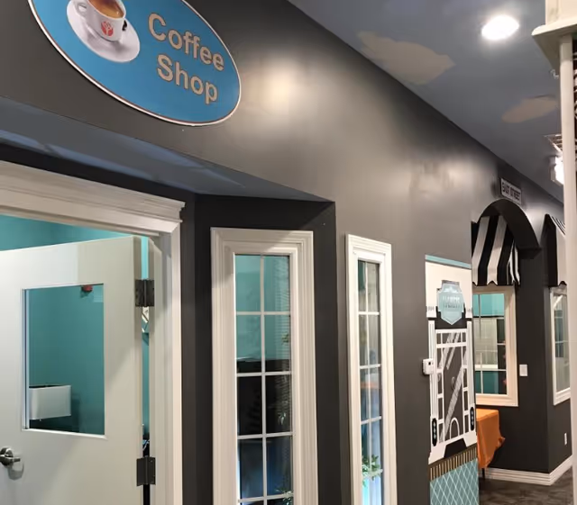 Interior hallway of a senior living facility with a coffee shop sign above an open door. The walls are painted gray with white trim around windows and doors. There is a decorative storefront facade with a black and white striped awning and a sign that reads 'Boutique'.