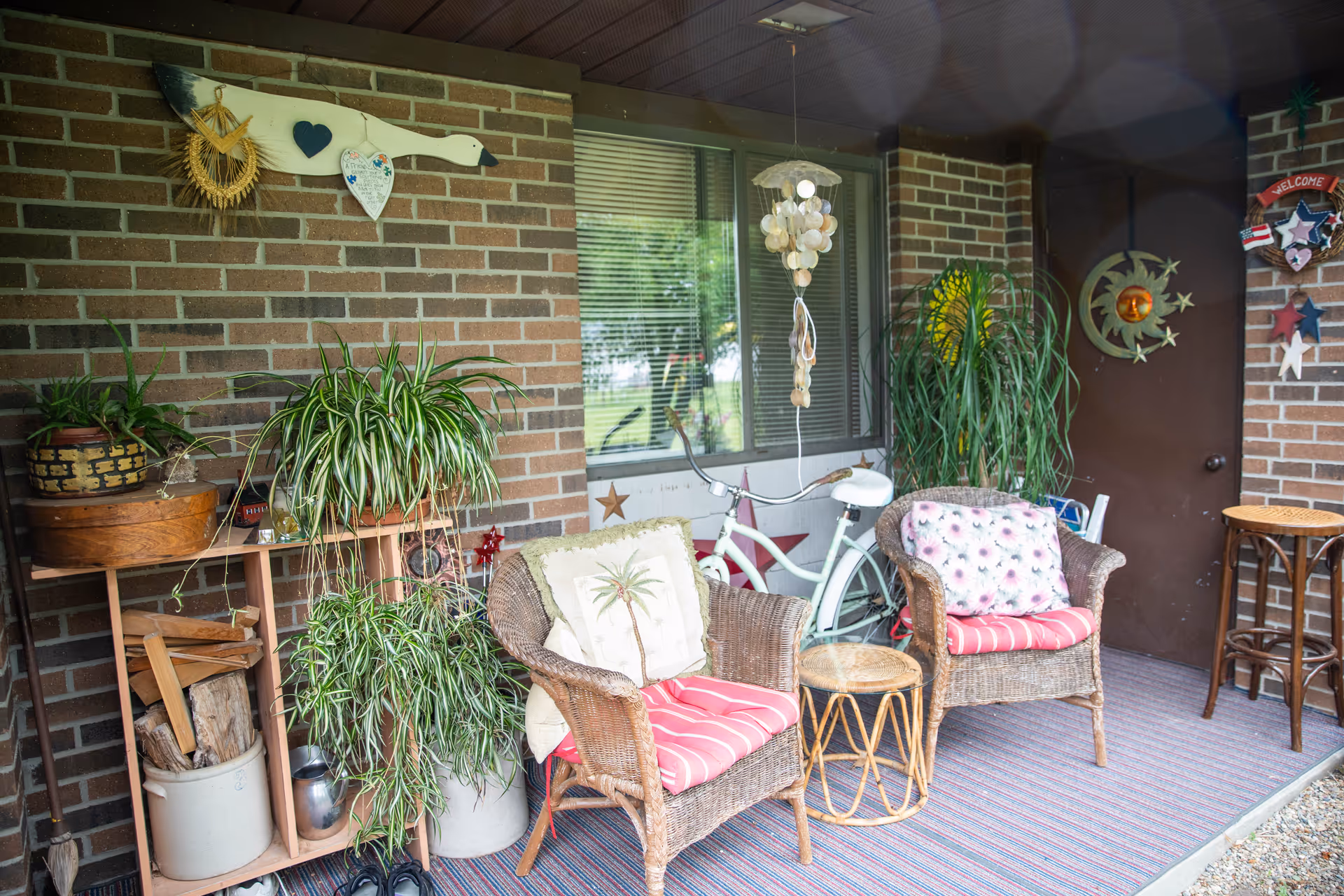 A cozy outdoor patio area with two wicker chairs featuring red and white cushions, a small round wicker table between them, various potted plants on a wooden shelf and on the floor, a white bicycle leaning against a brick wall, and decorative items including a hanging shell wind chime, a wooden duck wall decoration, and a sun and moon wall ornament near a brown door.