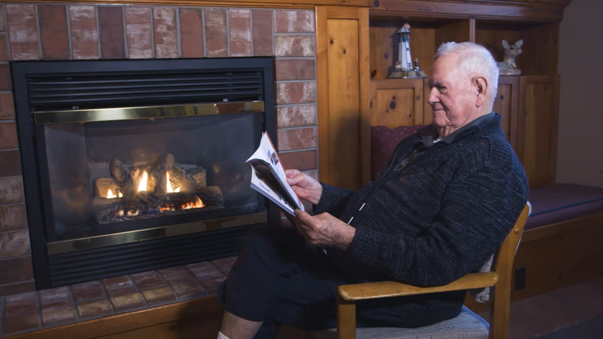 An elderly man sitting in a wooden chair reading a magazine in front of a lit brick fireplace inside a cozy room with wooden paneling and decorative items on shelves.