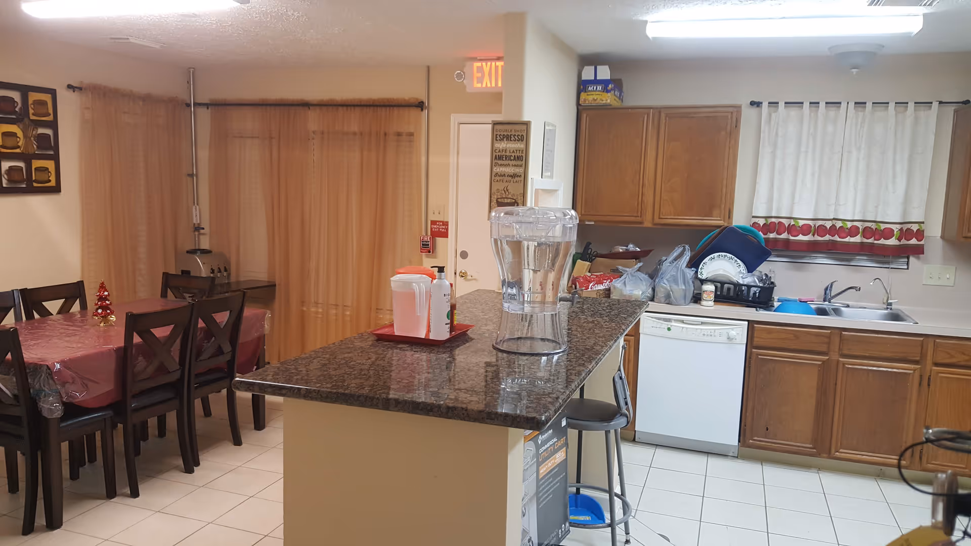 Interior view of a kitchen and dining area in an assisted living facility. The kitchen features wooden cabinets, a dishwasher, a sink with dishes drying on a rack, and a granite countertop island with a water dispenser and a pitcher. The dining area has a table covered with a red tablecloth and six chairs. Beige curtains cover a window and a door with an exit sign above it is visible in the background.