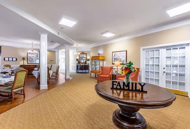Interior view of a senior living facility common area with a round wooden table in the foreground displaying a vase with flowers and a decorative 'FAMILY' sign. The room has beige walls, carpeted flooring, and several chairs and tables arranged for dining or socializing. There are white pillars, framed artwork on the walls, and a glass-paned door on the right side.