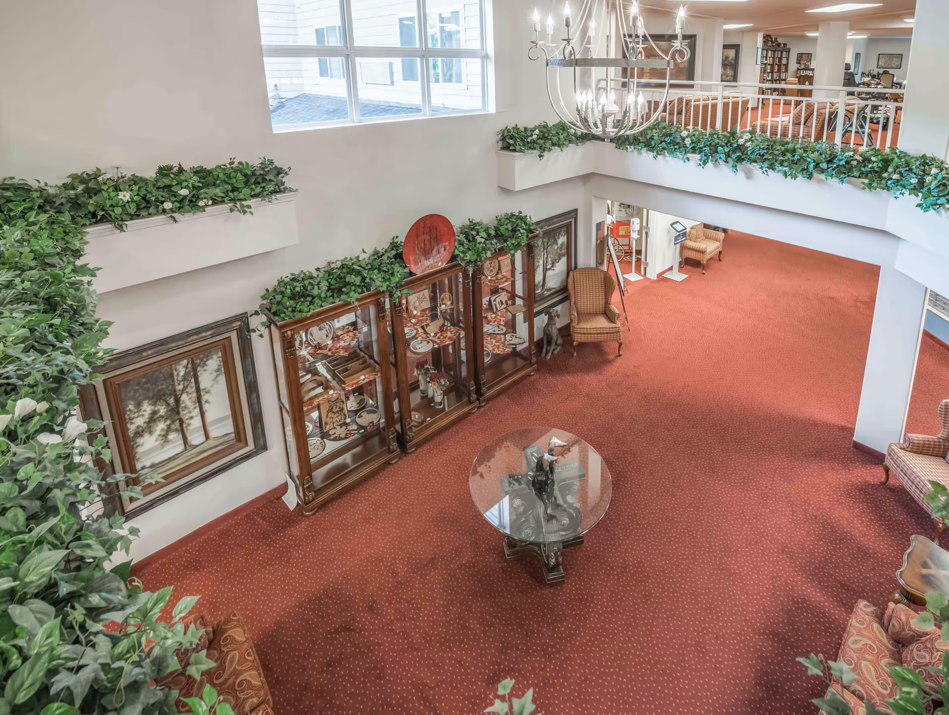 Interior view of a senior living facility lobby or common area with red carpet, a glass-top round table, display cabinets with decorative items, framed artwork on the walls, several upholstered chairs, green plants along the upper ledge, and a large chandelier hanging from the ceiling.