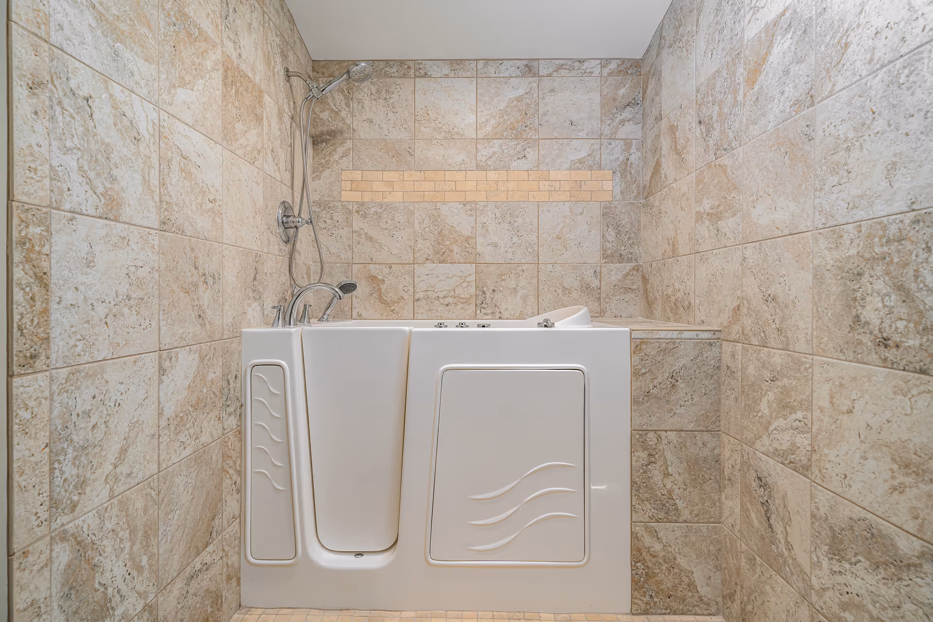 A walk-in bathtub with a side door for easy access, surrounded by beige and light brown tiled walls. The bathtub has a handheld showerhead and faucet fixtures on the left side.