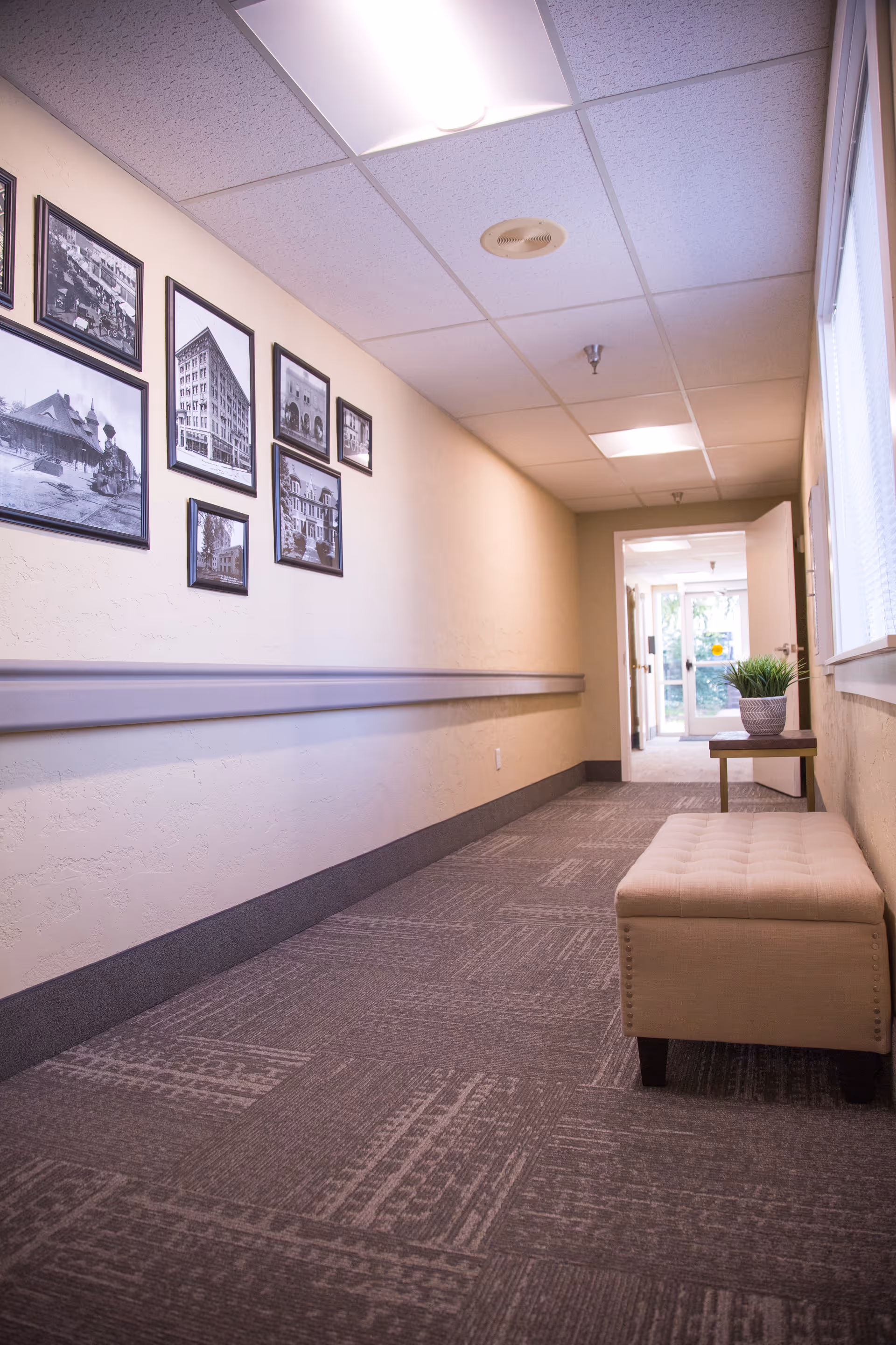 A well-lit hallway in a senior living facility with beige walls and carpeted floor. The left wall features several framed black and white photographs. On the right side, there is a cushioned bench and a small table with a potted plant near a window. The hallway leads to a glass door at the end, allowing natural light to enter.