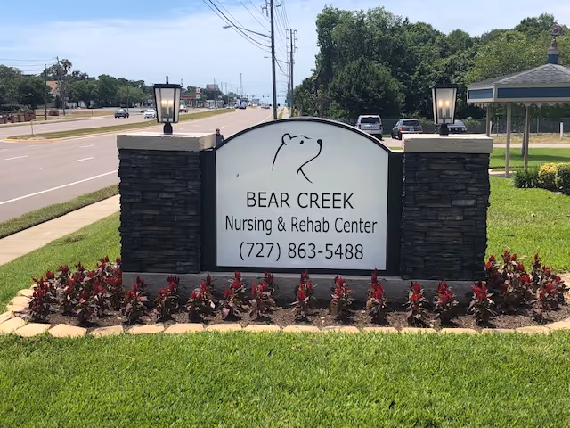 Outdoor sign for Bear Creek Nursing & Rehab Center with a bear logo, phone number, and two stone pillars with lamps on each side, surrounded by red plants and green grass, next to a road with cars and trees in the background.