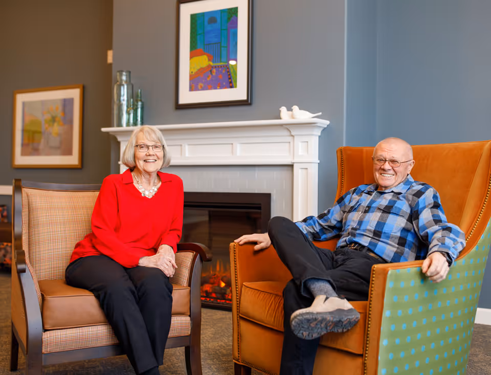 Two older adults smiling while seated in armchairs in a cozy lounge area with a fireplace and framed artwork.
