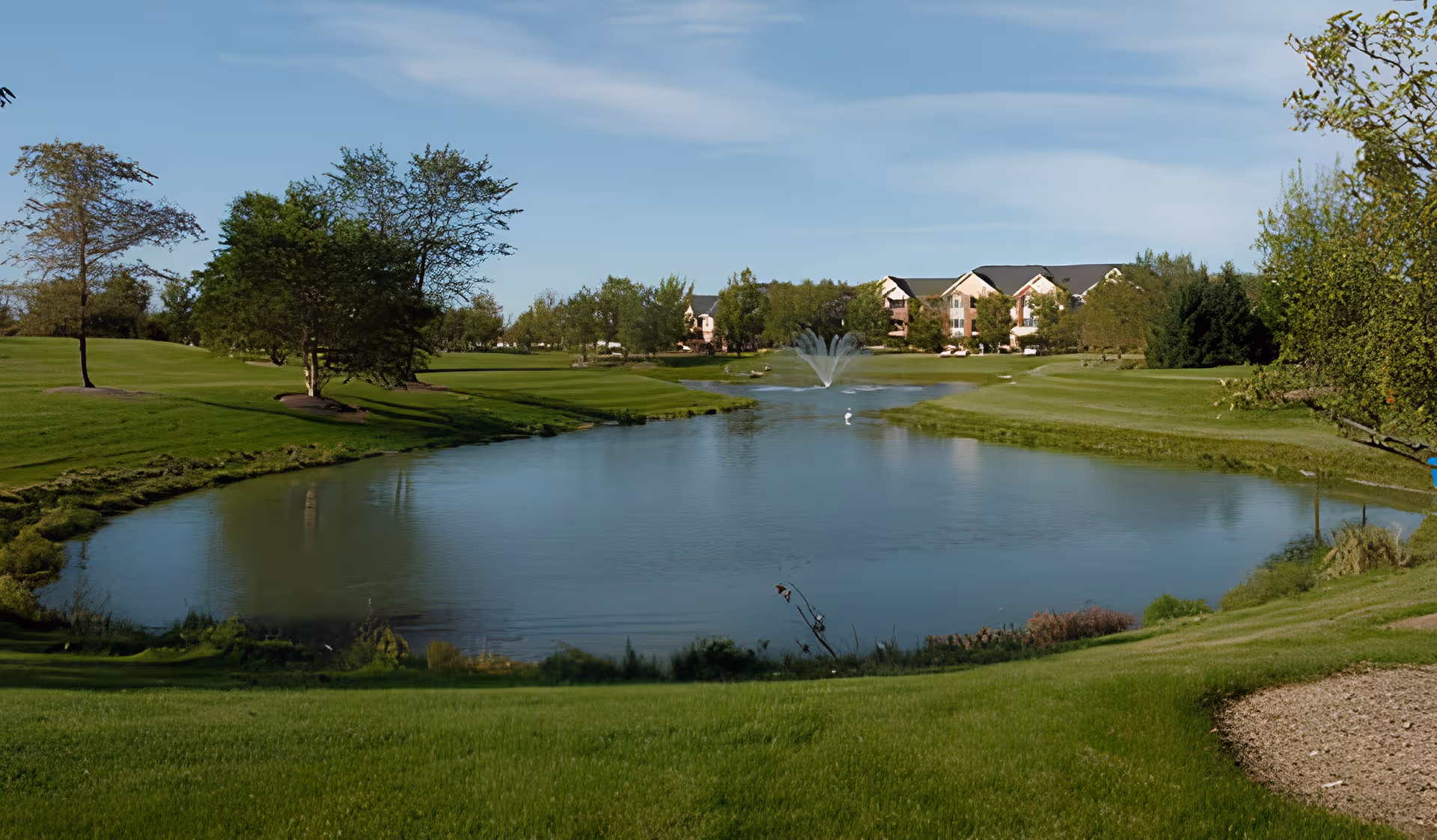 A scenic outdoor view of a pond with a water fountain in the center, surrounded by well-maintained green grass and trees. In the background, there are buildings partially visible behind the trees under a blue sky with some clouds.