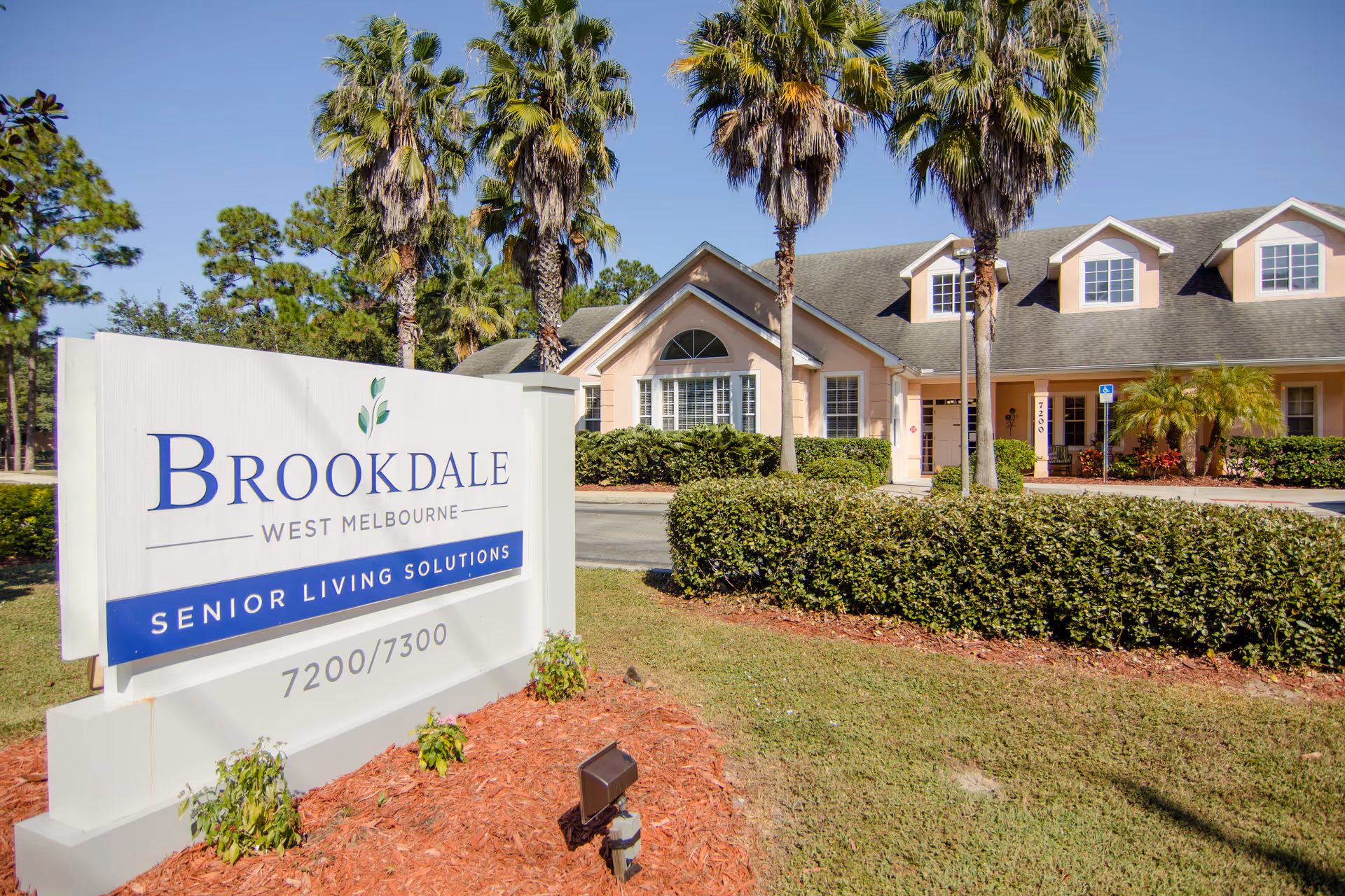 Exterior view of Brookdale West Melbourne senior living facility with a large white sign in the foreground displaying the facility name and address. The building is light peach with white trim, surrounded by palm trees, bushes, and a well-maintained lawn under a clear blue sky.
