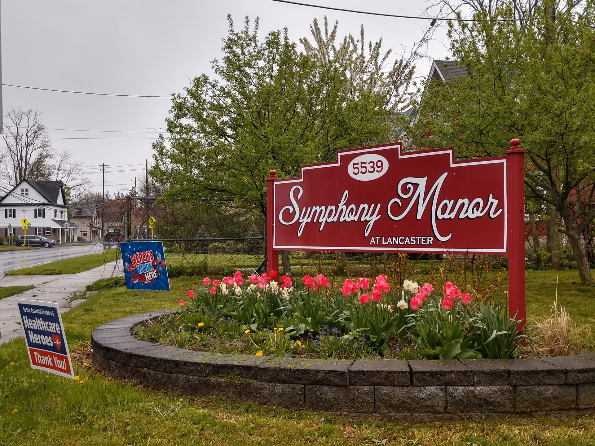 Outdoor view of a red sign for Symphony Manor at Lancaster with the address number 5539. The sign is surrounded by a flower bed with blooming tulips and other flowers. There are trees and a sidewalk nearby, with a couple of smaller signs expressing thanks to healthcare heroes.