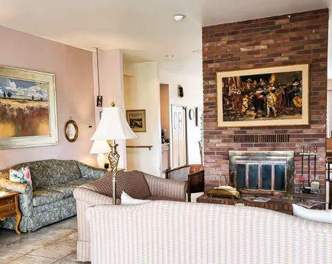 A cozy living room with a brick fireplace, a painting above the fireplace, two sofas with patterned upholstery, a floor lamp, and framed artwork on the walls.