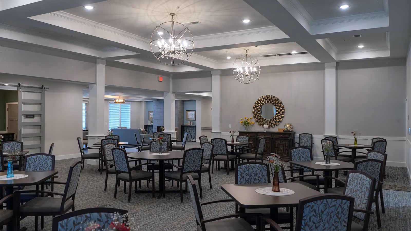 A spacious dining room with multiple square and round tables surrounded by chairs with patterned upholstery. Each table has a small vase with flowers. The room features modern chandeliers hanging from a coffered ceiling, neutral-colored walls, and a decorative mirror above a dark wooden sideboard. In the background, there is a seating area with sofas and large windows with blinds.