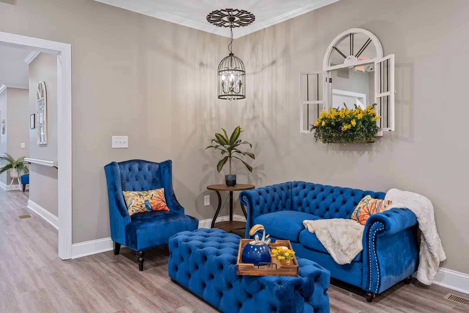 A cozy sitting area in a senior living facility featuring a blue velvet tufted sofa and matching armchair with colorful cushions. A tufted blue ottoman with a wooden tray holding a blue teapot and a small yellow flower arrangement is in front of the sofa. A small round wooden side table with a potted plant is placed between the sofa and chair. The walls are painted beige with a decorative hanging light fixture above and a wall-mounted window frame with yellow flowers inside.