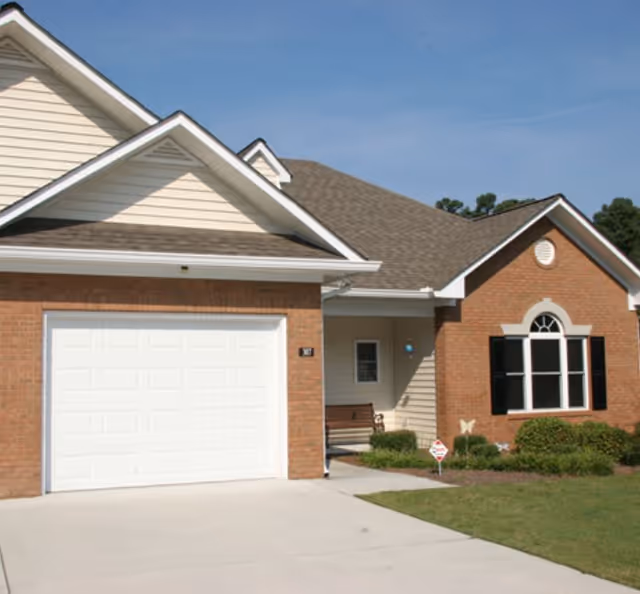 Exterior view of a single-story residential building with a brick and beige siding facade, a white garage door, a small porch with a bench, and a well-maintained lawn under a clear blue sky.