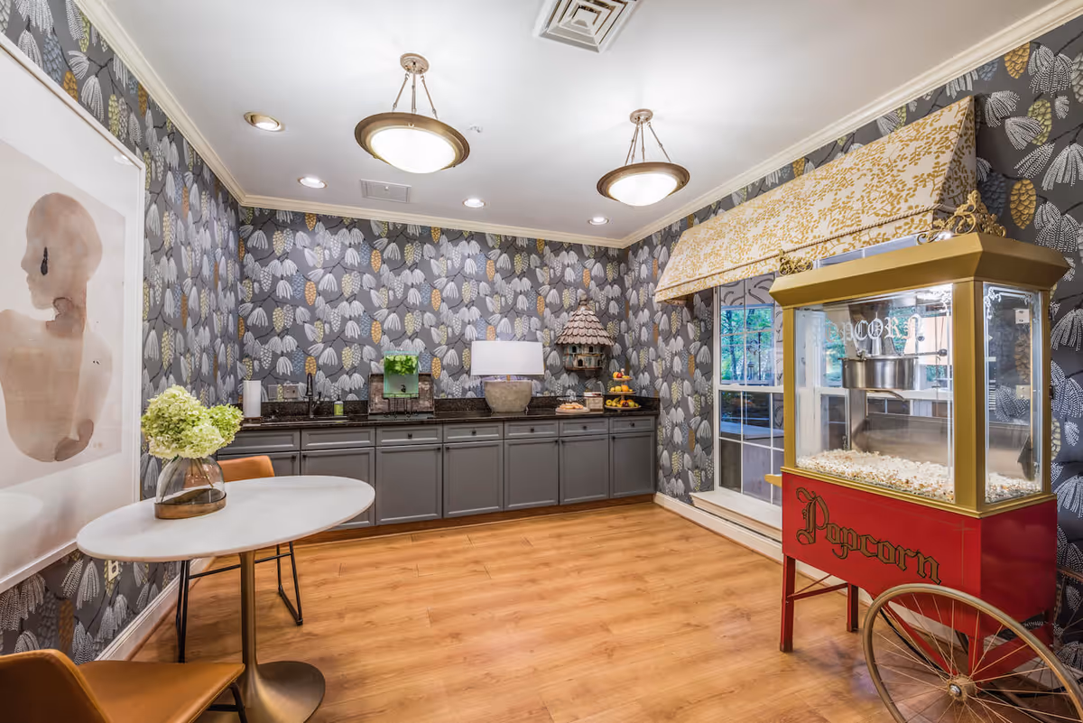 A cozy interior room with floral patterned wallpaper, wooden flooring, and a popcorn machine on the right side. There is a small round table with two brown chairs and a vase with green flowers on the left. The back wall features gray cabinets with a countertop holding a lamp, a birdhouse, a fruit stand, and other decorative items. A window with a patterned valance is also visible.