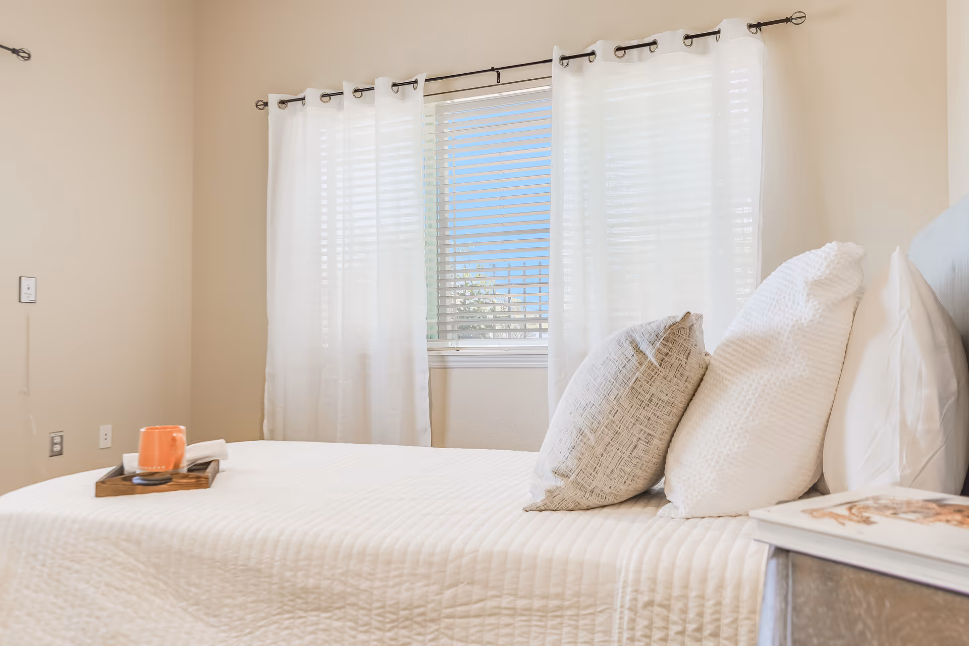 Bright bedroom with a made bed, decorative pillows, a tray with a mug, and a window with white curtains.