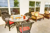 Outdoor patio area with a round glass table surrounded by four wicker chairs with red cushions. On the table are plates with snacks and glasses of iced tea. In the background, there are cushioned wooden chairs and a loveseat with a yellow pillow that says 'our nest'. The patio is adjacent to a building with white siding and several windows.
