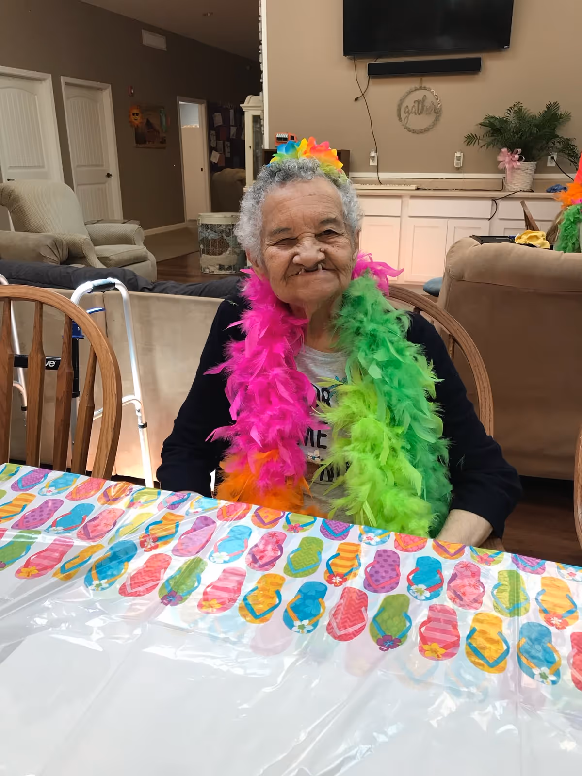 An elderly woman sitting at a table covered with a colorful flip-flop patterned tablecloth. She is wearing bright pink and green feather boas and a multicolored flower headband. The background shows a cozy living room area with chairs, a walker, a TV mounted on the wall, and some plants.