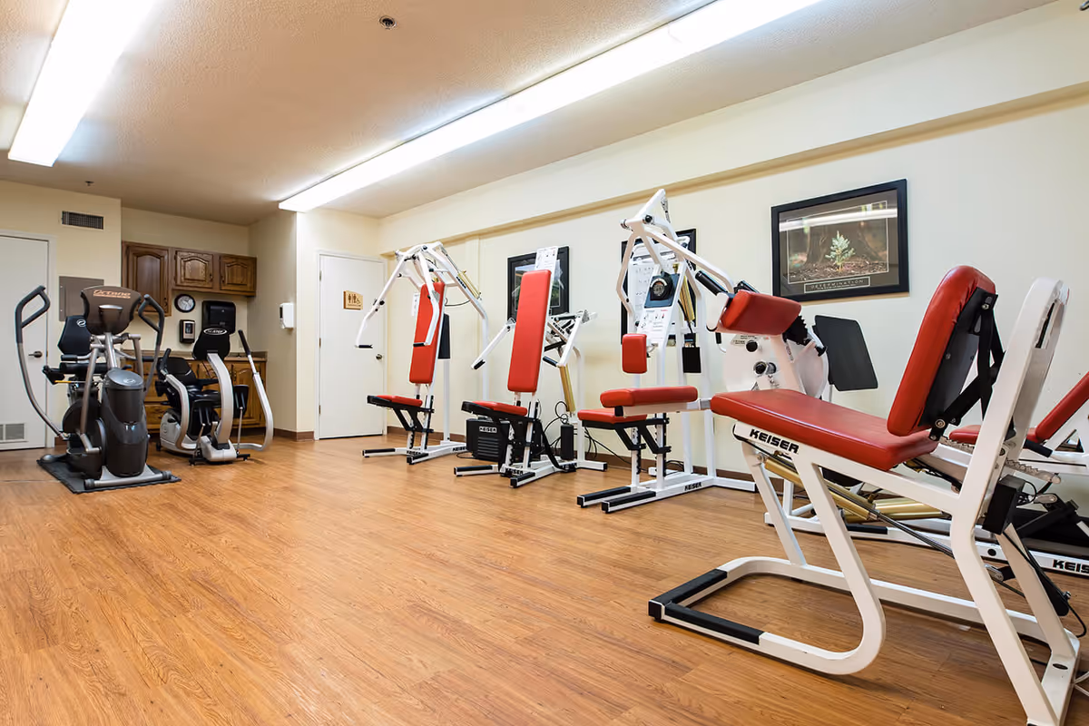 A fitness room in an assisted living facility with various exercise machines including elliptical trainers and strength training equipment with red padded seats and white frames. The room has wood flooring, fluorescent ceiling lights, and motivational framed pictures on the walls.