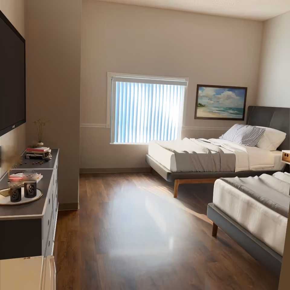 A bright bedroom with two beds featuring gray upholstered headboards and white bedding. A large window with vertical blinds lets in natural light. A dresser with decorative items and a flat-screen TV mounted above it is on the left side. A framed painting of a beach scene hangs on the wall above one of the beds. The room has wooden flooring and neutral-colored walls.