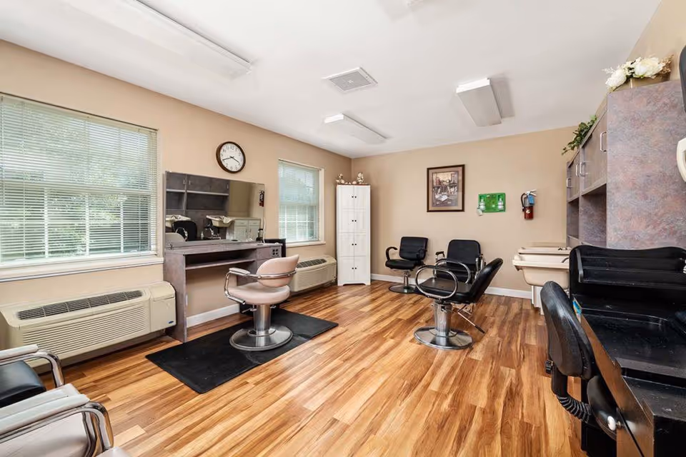 A bright room with wooden flooring featuring salon chairs, mirrors, and hair washing sinks, likely a hair salon or grooming area within a facility. The room has two windows with blinds, a clock on the wall, and various cabinets and shelves.