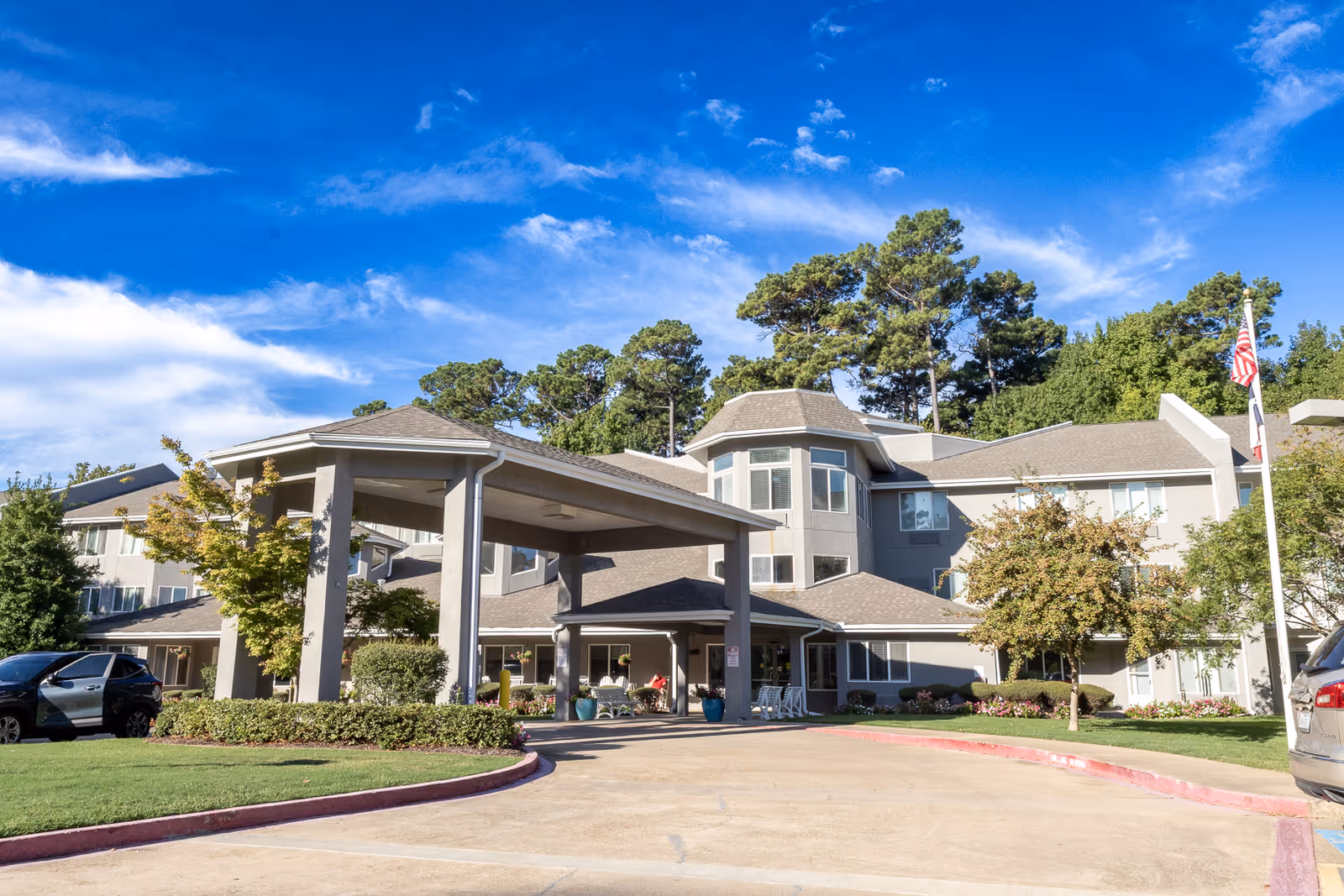Exterior front entrance of a multi-story senior living facility with a covered porte-cochère, flagpole, parked cars, and landscaped grounds under a blue sky.