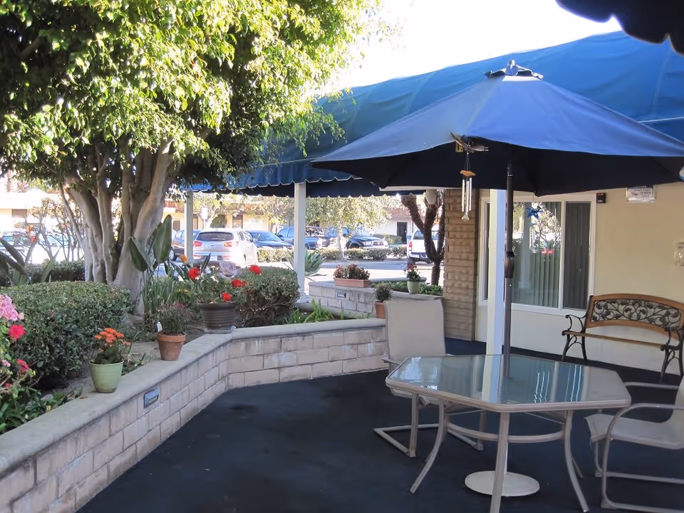 Shaded outdoor patio with a glass table, chairs, an umbrella, potted plants, and a bench by the building entrance.