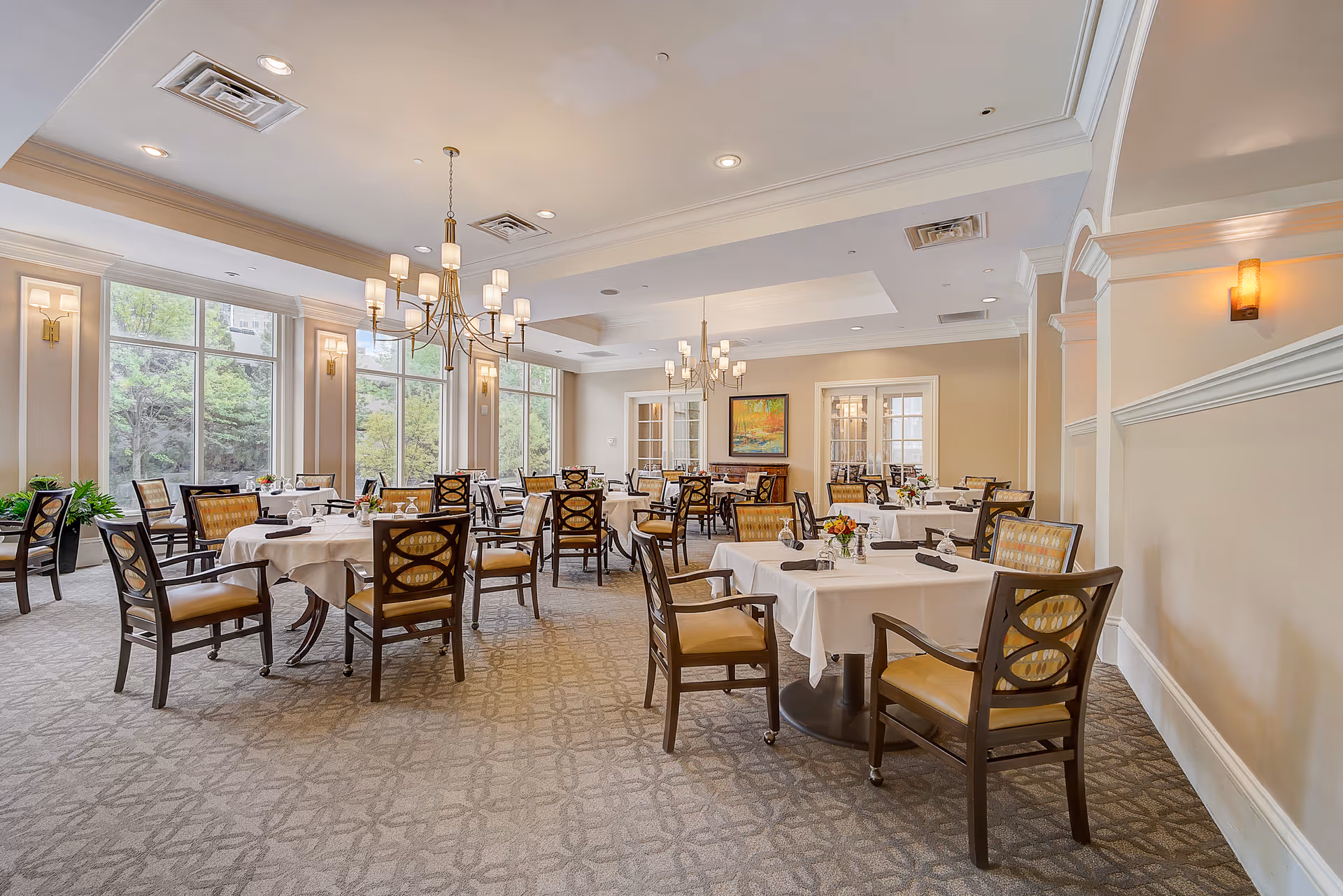 Bright dining room with tables covered in white linens, wooden chairs, chandeliers, and large windows overlooking greenery.