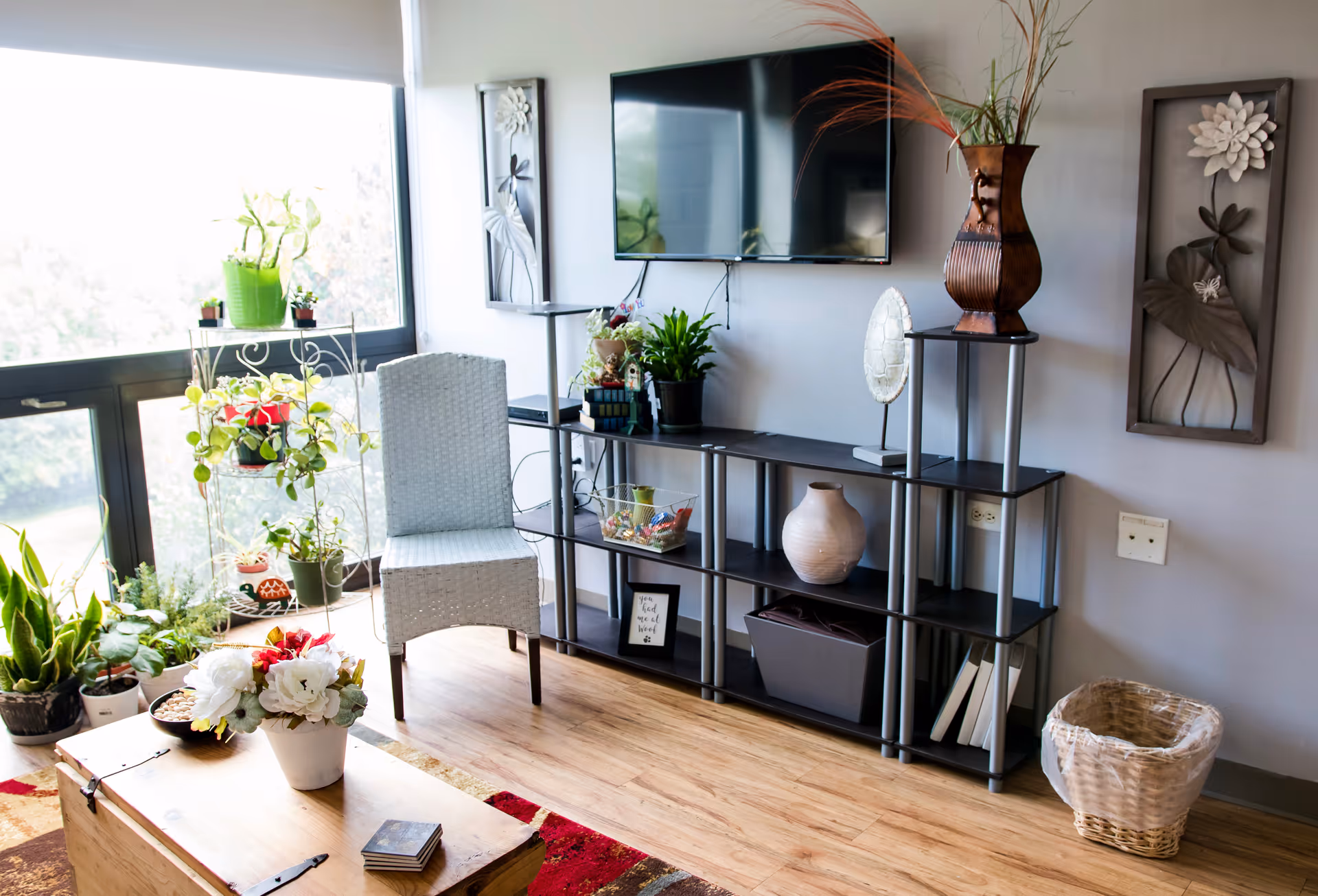 A bright living room area with a large window letting in natural light. There is a white wicker chair next to a black shelving unit holding various decorative items including plants, vases, and framed art. A flat-screen TV is mounted on the wall above the shelving unit. A wooden coffee table with a flower arrangement sits on a colorful rug in the foreground. The room has wooden flooring and light gray walls with floral wall art and a wicker basket on the floor.