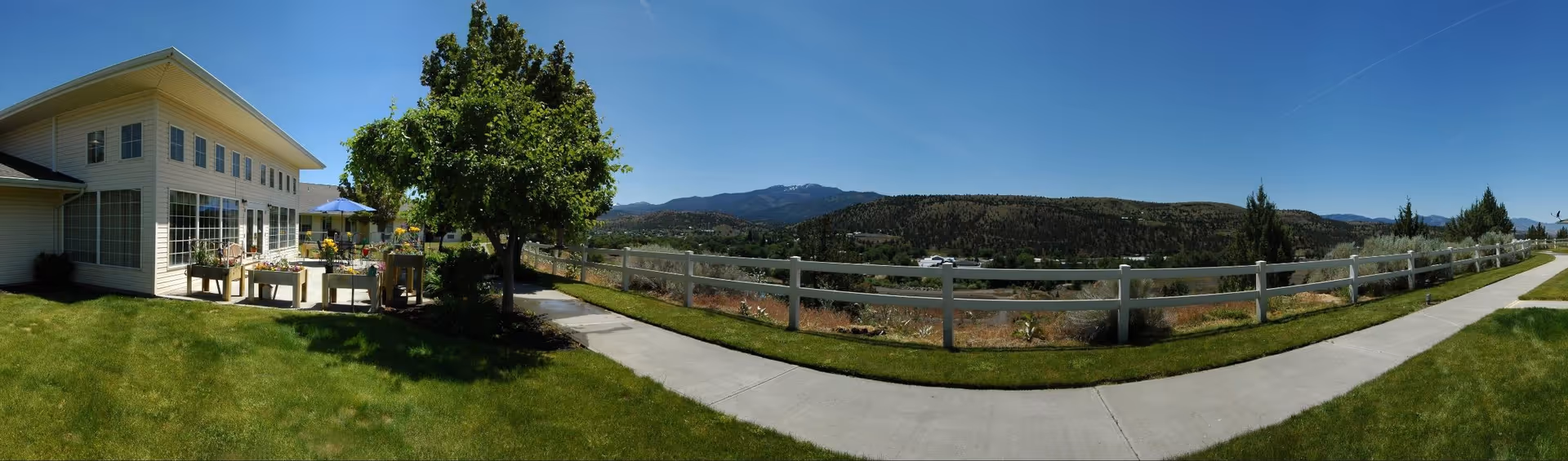 Outdoor view of a senior living facility named Sapphire at Valley View showing a white building with large windows, a patio area with tables and umbrellas, a tree, a white fence, a sidewalk, green grass, and a scenic background of hills and mountains under a clear blue sky.
