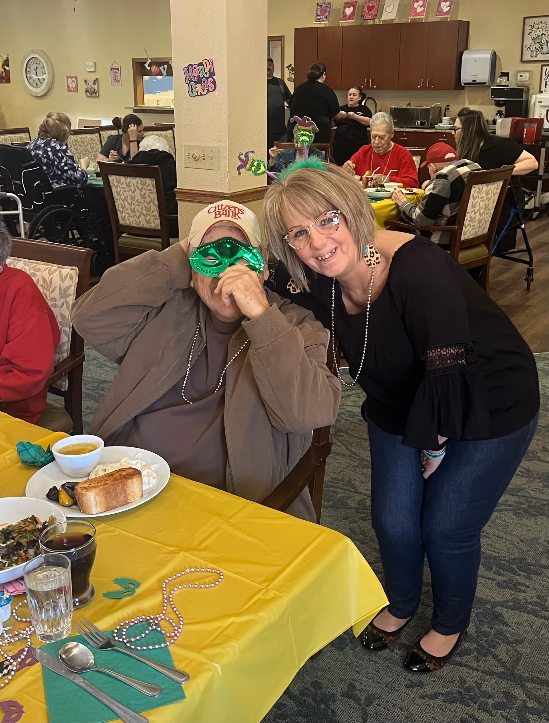 A festive dining room scene at Kyrie Assisted Living with residents and staff enjoying a meal. A man wearing a Citizens Bank cap and a green Mardi Gras mask sits at a table with a yellow tablecloth, plates of food, and drinks. A woman with glasses and a black top leans in smiling next to him. Other residents and staff are visible in the background, some seated and eating, others standing and conversing. Mardi Gras decorations are visible on the walls and table.