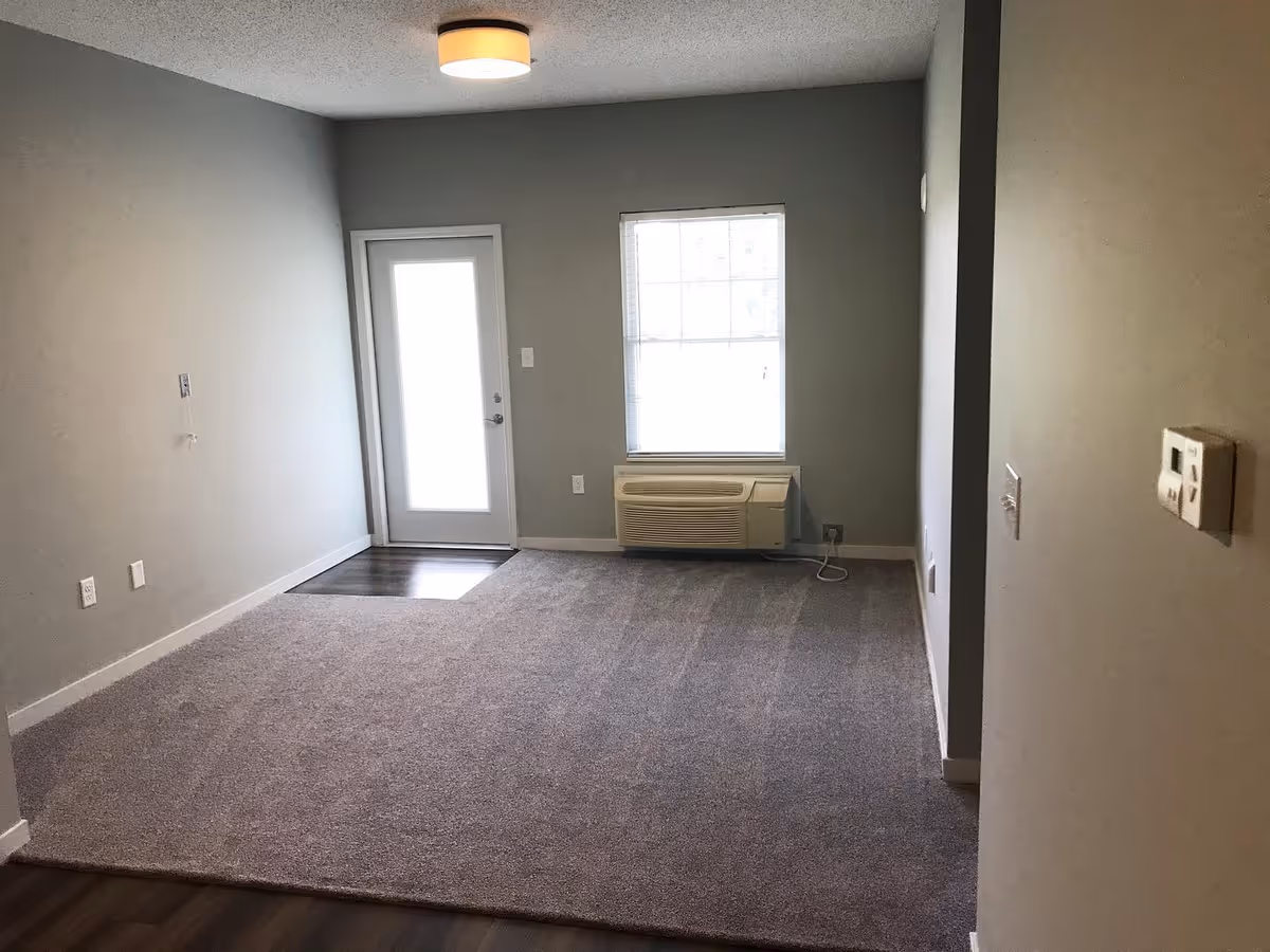 Empty living room with carpeted floor, a window with a wall air-conditioning unit, and a door letting in light.