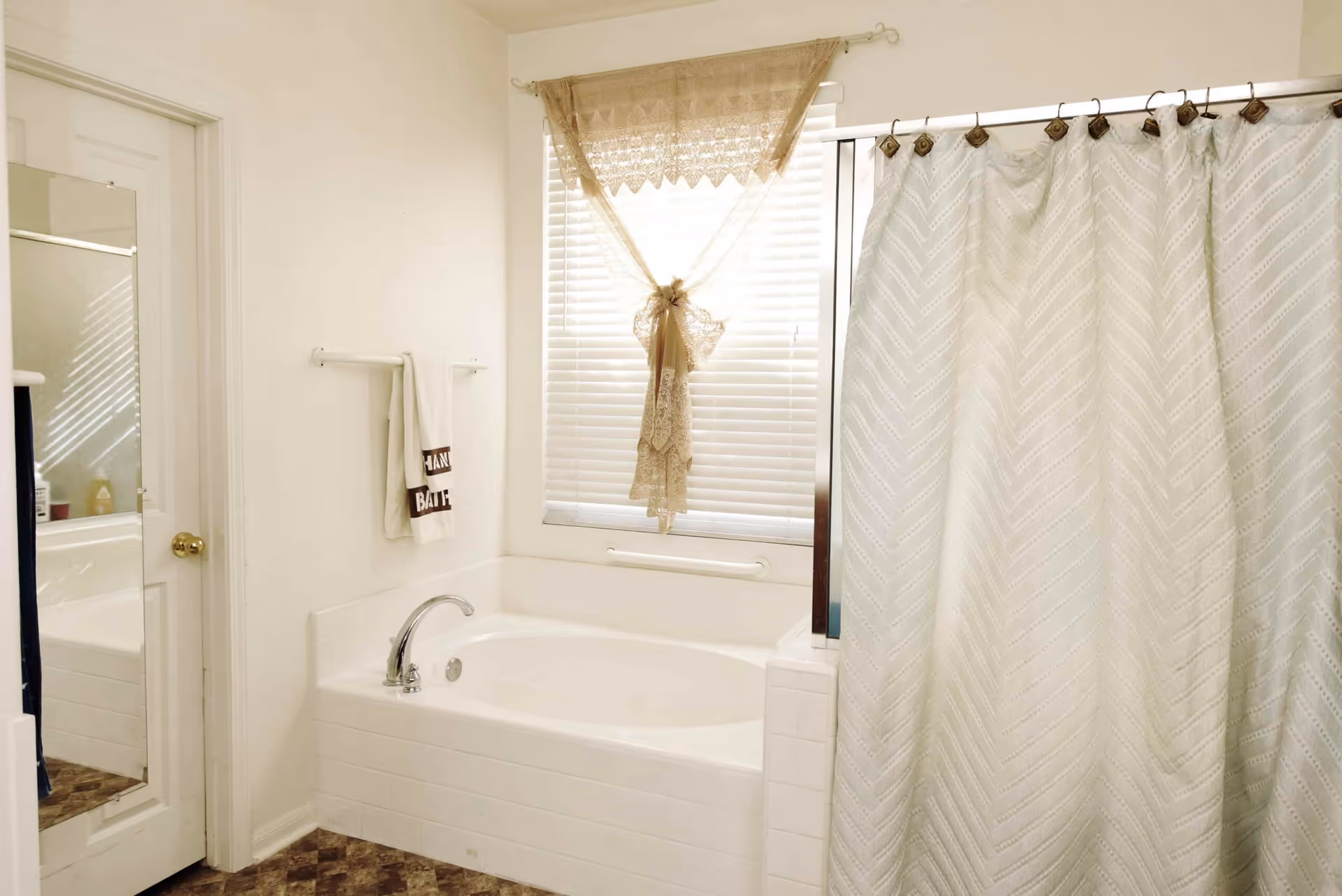 A bright bathroom featuring a white bathtub with a silver faucet beneath a window covered with white blinds and a decorative beige lace curtain tied in the center. A white towel hangs on a wall-mounted towel rack next to the tub. A door with a full-length mirror is visible on the left side, and a white shower curtain with a subtle chevron pattern is partially drawn on the right side.