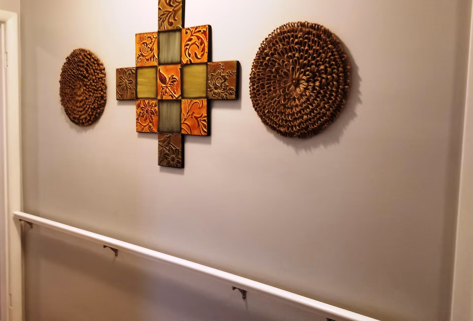 A hallway wall decorated with three pieces of wall art: a square arrangement of colorful tiles with floral patterns in the center, flanked by two round, textured brown decorative pieces. A white handrail runs along the wall below the artwork.