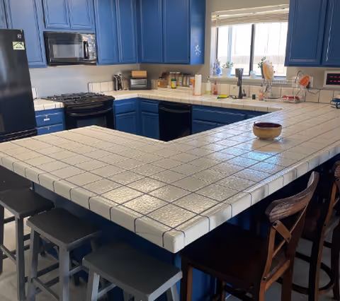 A kitchen with blue cabinets and white tiled countertops. The kitchen features a black refrigerator, a microwave, a stove, a dishwasher, and a sink under a window. There are several stools around the countertop, and various kitchen items such as paper towels, spices, and a bowl are visible on the counter.