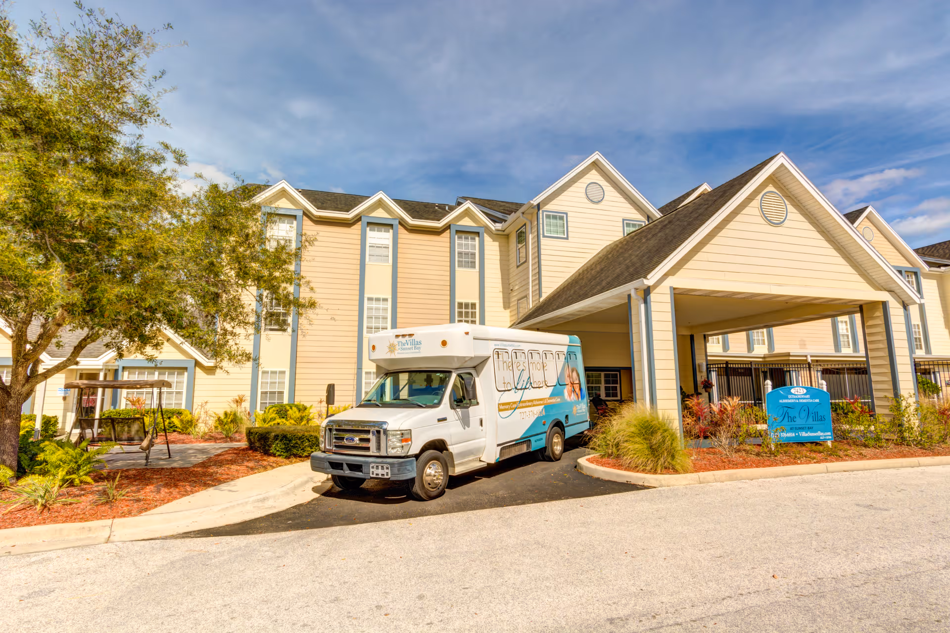Exterior front entrance of a beige multi-story senior living facility with a shuttle van parked under the porte-cochère.