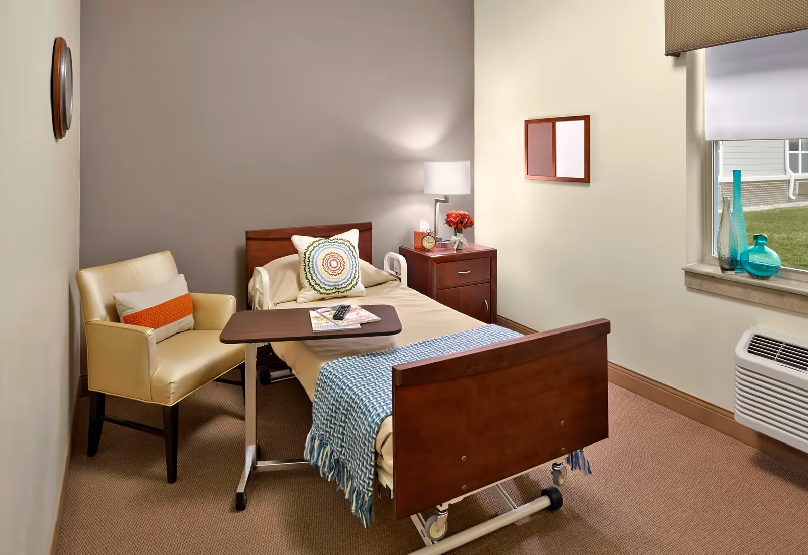 A neatly arranged senior bedroom with a single adjustable wooden bed, bedside table and lamp, armchair, overbed table, and a window with decorative vases.