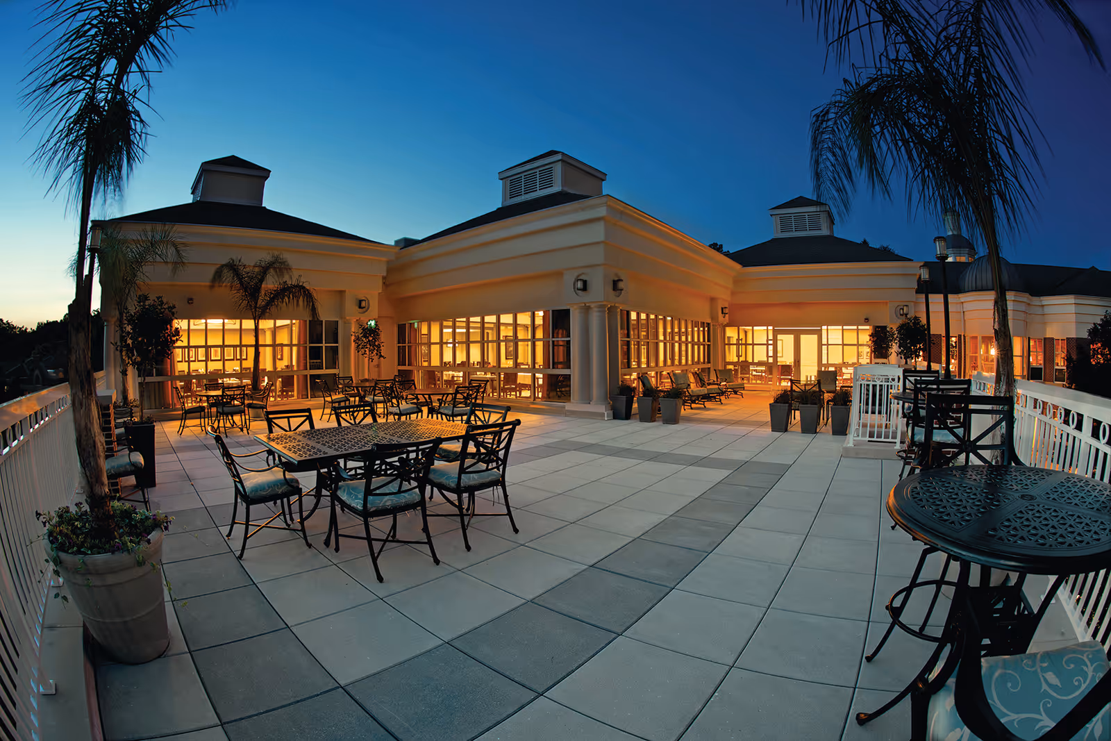 Outdoor patio area at dusk with multiple tables and chairs arranged on a tiled floor, surrounded by potted palm trees and plants. The building in the background has large windows with warm interior lighting visible.